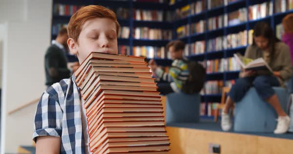 Preteen boy carrying pile of heavy books in school library ...