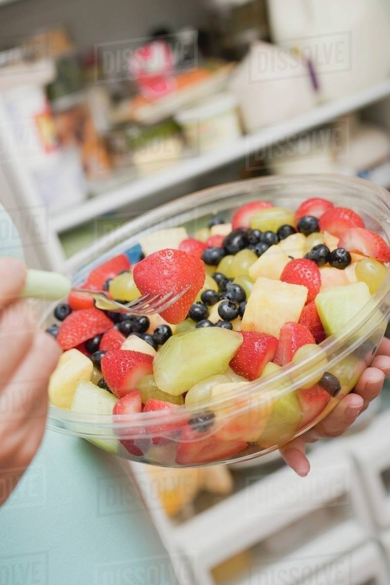 Woman eating fruit salad out of plastic bowl in front of refrigerator -  Royalty-free Stock Photo | Dissolve