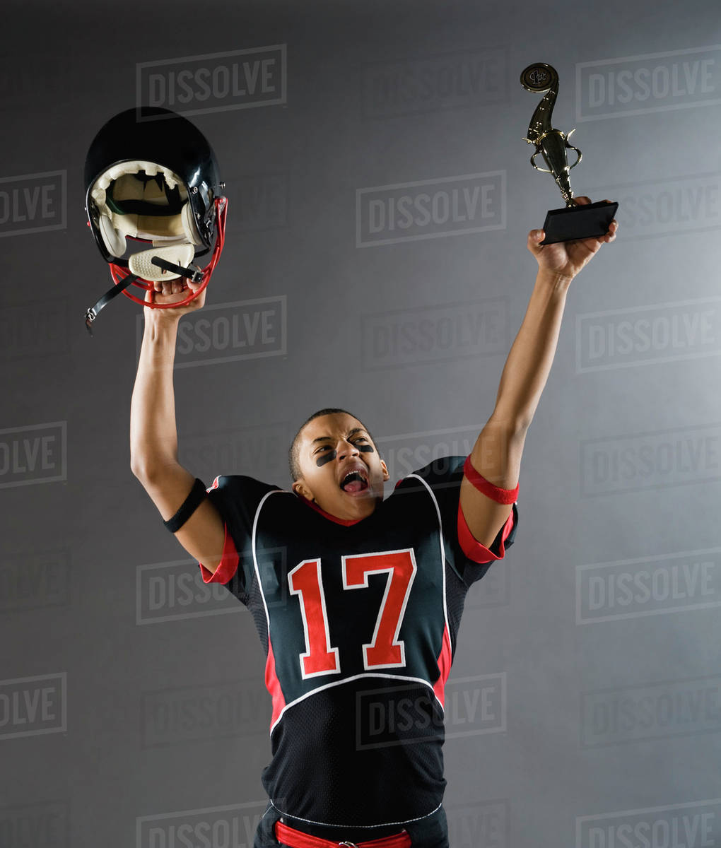 Mixed race football player holding helmet and trophy overhead Stock