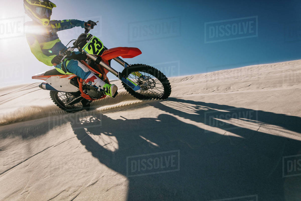 Motocross biker riding his racing bike on a sand dune with his shadow in front. Motorbike rider in racing gear riding up the sand dune leaving a trail of sand dust with sun in the background. Royalty-free stock photo