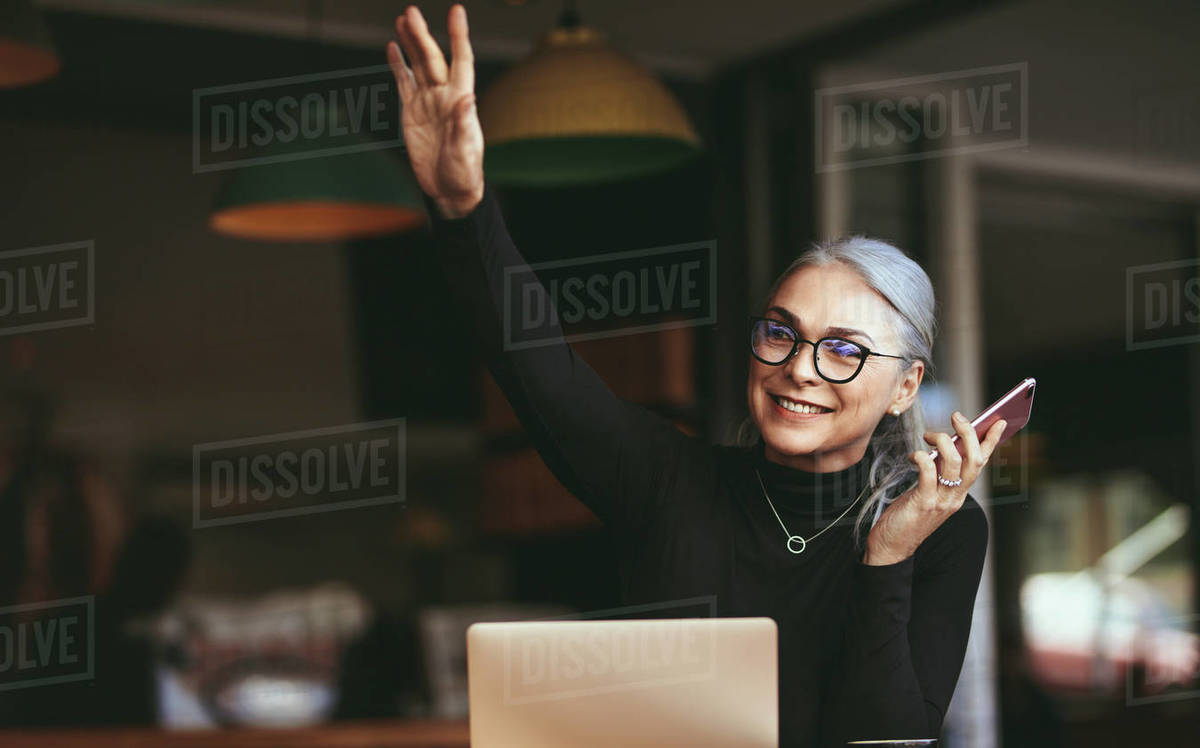 Portrait of happy senior woman sitting at cafe with laptop talking on mobile phone and waving at someone. Mature businesswoman at coffee shop. Royalty-free stock photo