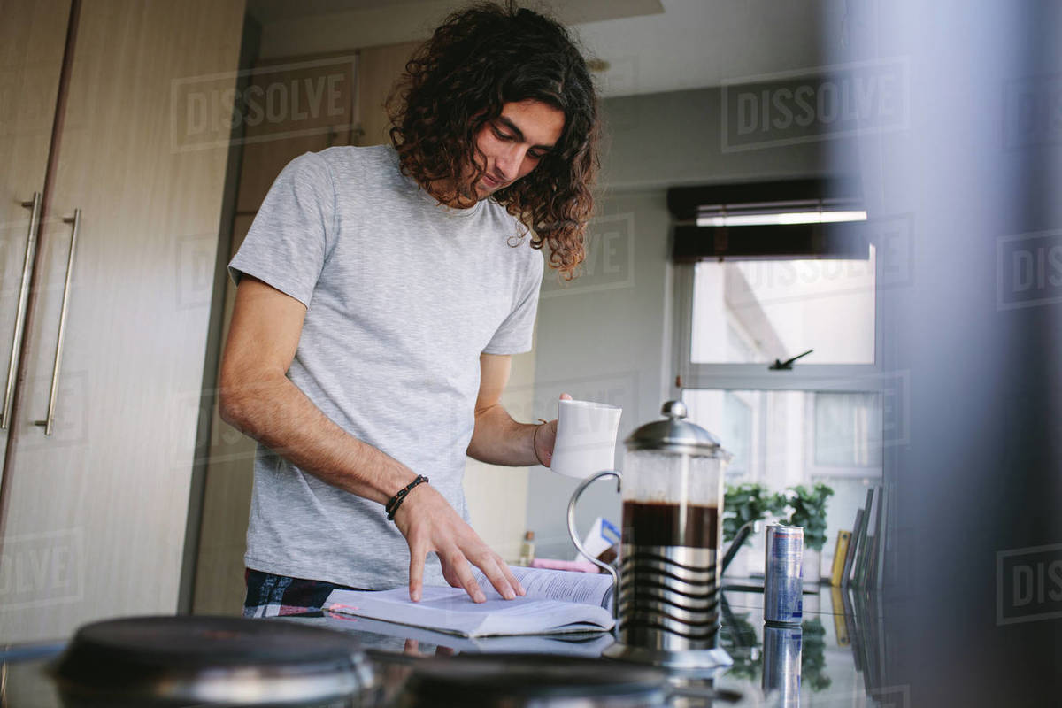 Student reading a book in kitchen while drinking coffee. Smiling