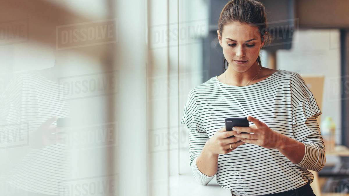 Working woman standing beside a window holding mobile phone. Woman entrepreneur taking a break from work checking her cell phone. Royalty-free stock photo