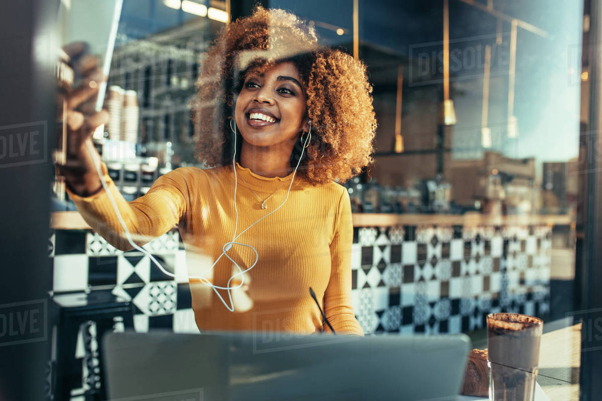 Woman freelancer sitting in a coffee shop taking a selfie on cell phone with a laptop in front. Smiling afro american woman doing a video call on mobile phone sitting at a restaurant. Royalty-free stock photo