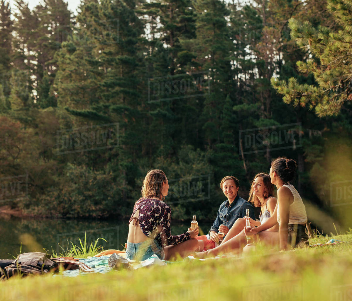 Group of teenagers having a picnic on the lake. Young friends sitting together with beers near a