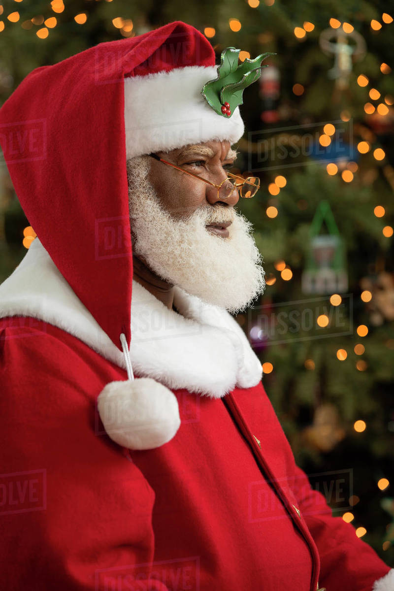 An African American man dressed as Santa Claus sitting in front of a Christmas tree turned profile, looking off camera. Royalty-free stock photo