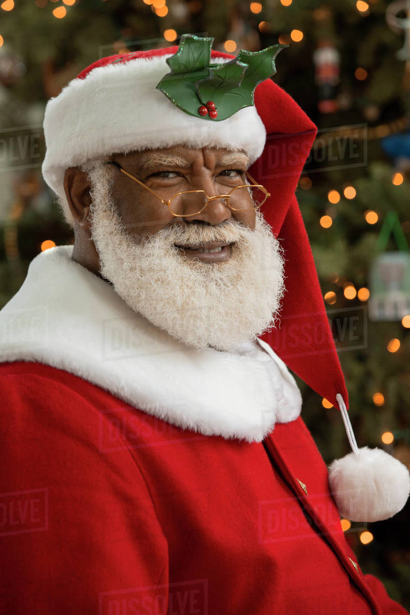 An African American man dressed as Santa Claus sitting in front of a Christmas tree looking camera smiling.  Royalty-free stock photo