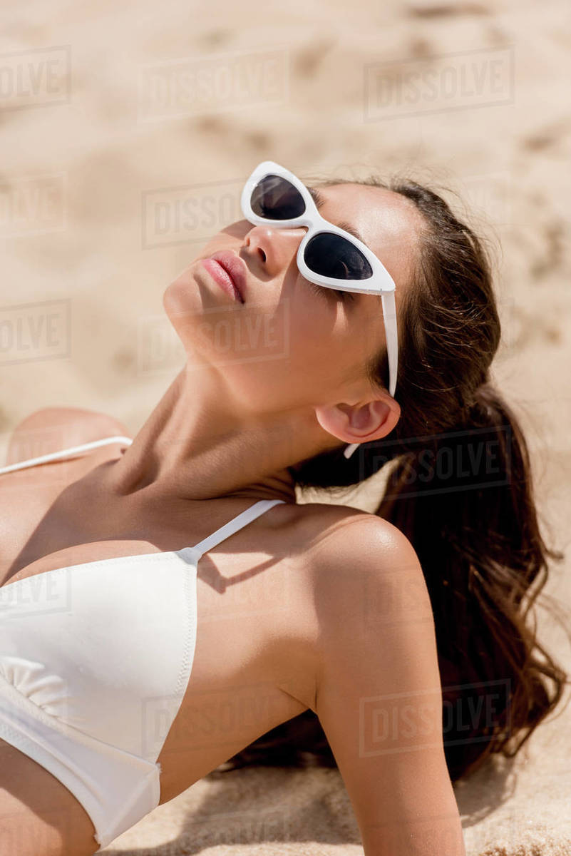 beautiful young woman in sunglasses and white bikini sunbathing on sand