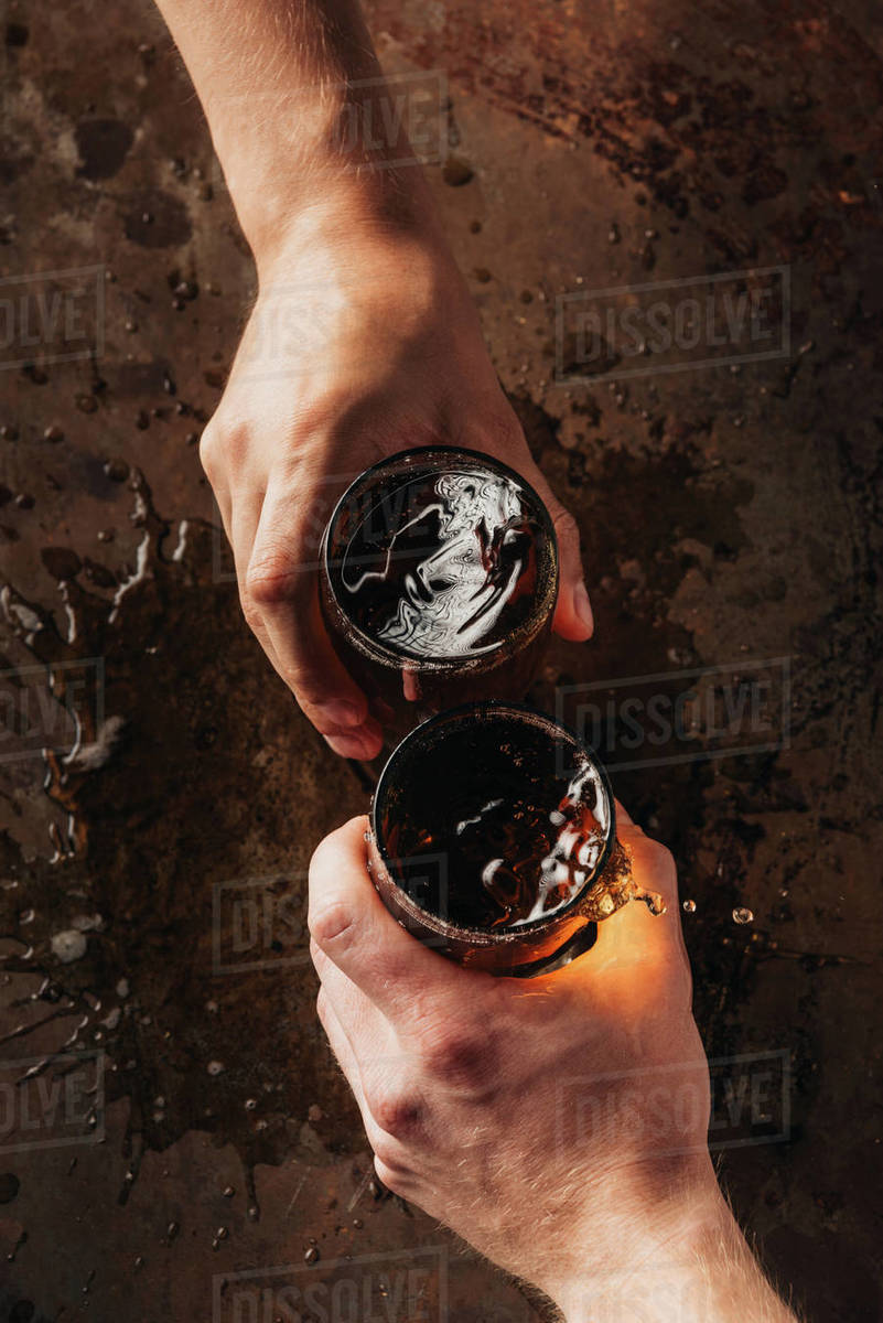 cropped shot of friends clinking mugs of beer at dark surface Stock