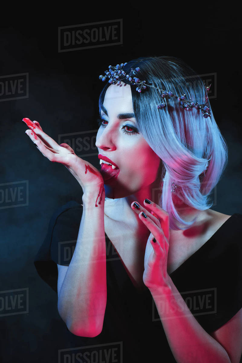 Attractive vampire licking blood from hand on dark background with
