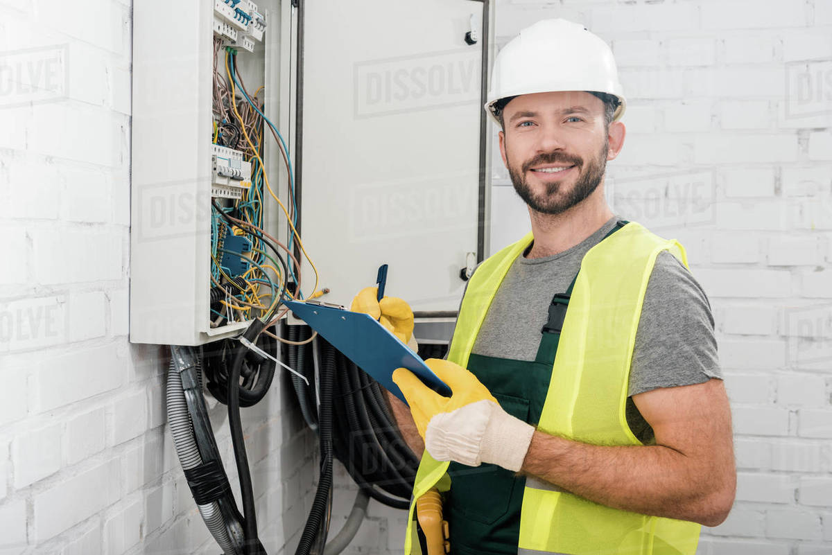 Services Smiling handsome electrician holding clipboard near electrical box in corridor and looking at camera - Royalty-free Stock Photo | Dissolve