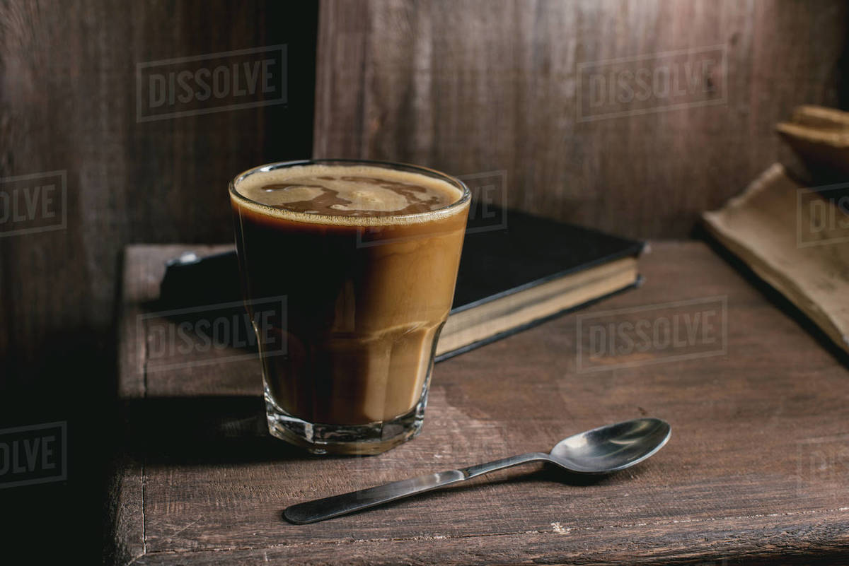 Glass of coffee drink, old book and spoon on rustic wooden table near