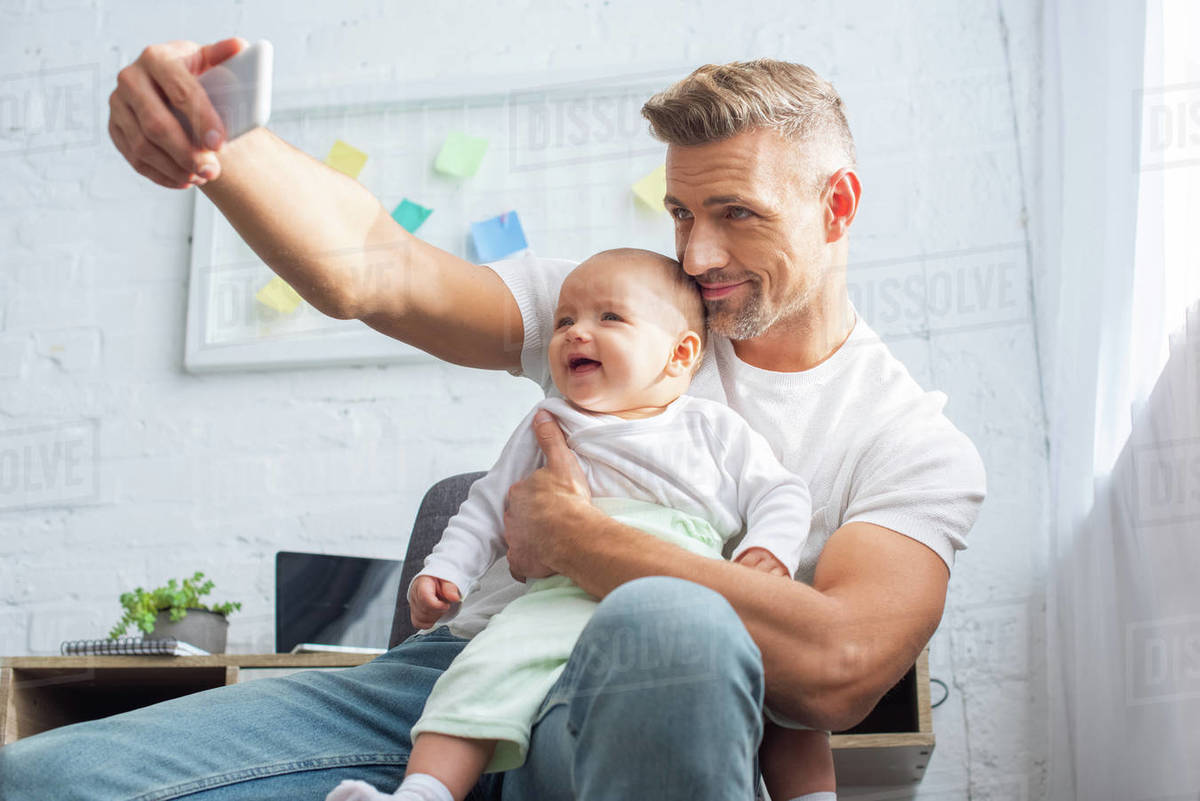 Happy father sitting on chair, holding baby daughter and taking selfie