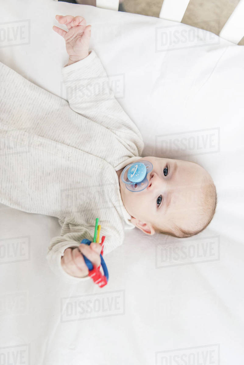 Overhead view of little baby with pacifier holding toy while lying in