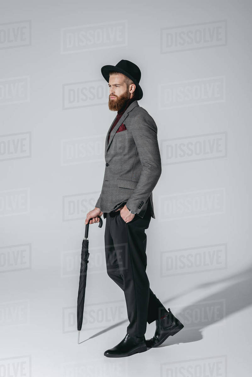 side view of stylish man in hat holding umbrella isolated on grey