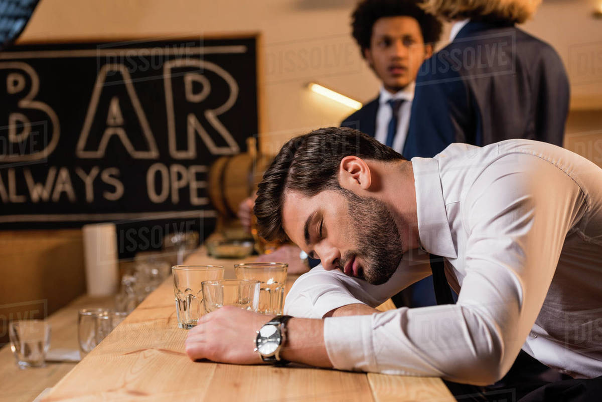Young drunk businessman sleeping on bar counter while colleagues