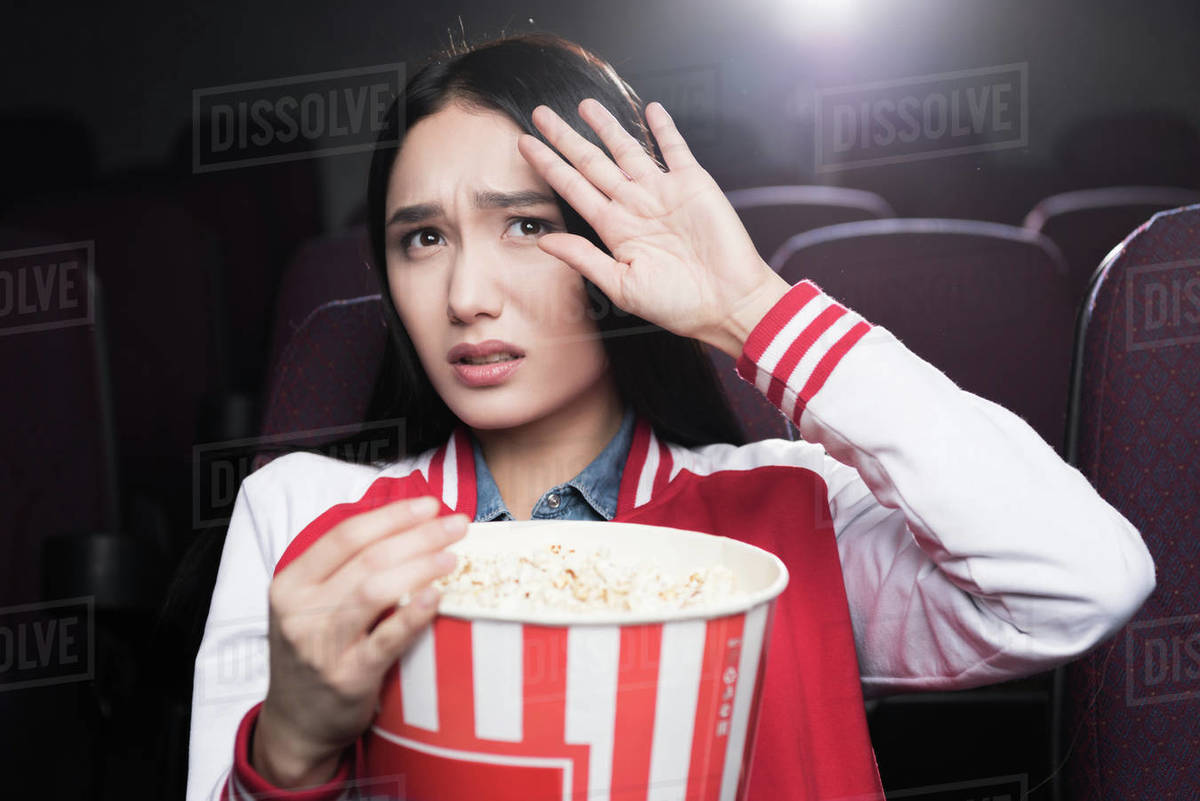 frightened asian girl eating popcorn and watching movie in cinema