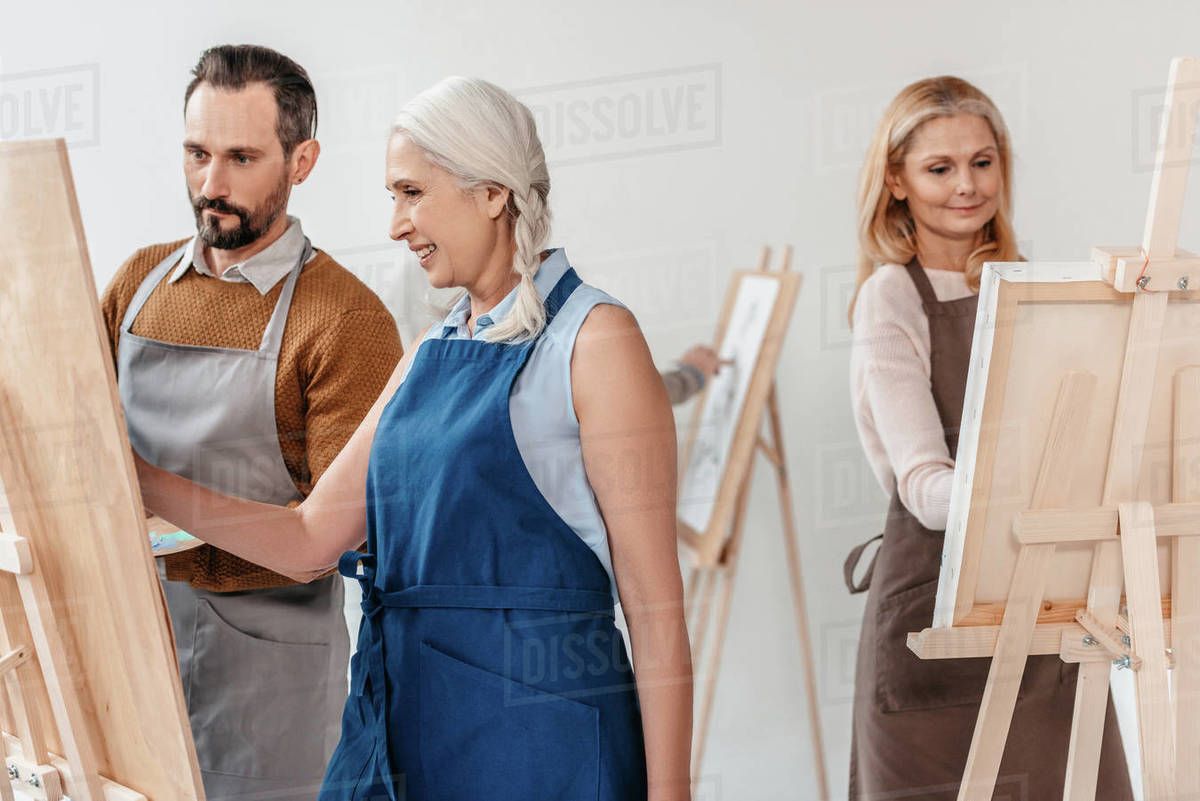 mature students in aprons painting on easels during art class for
