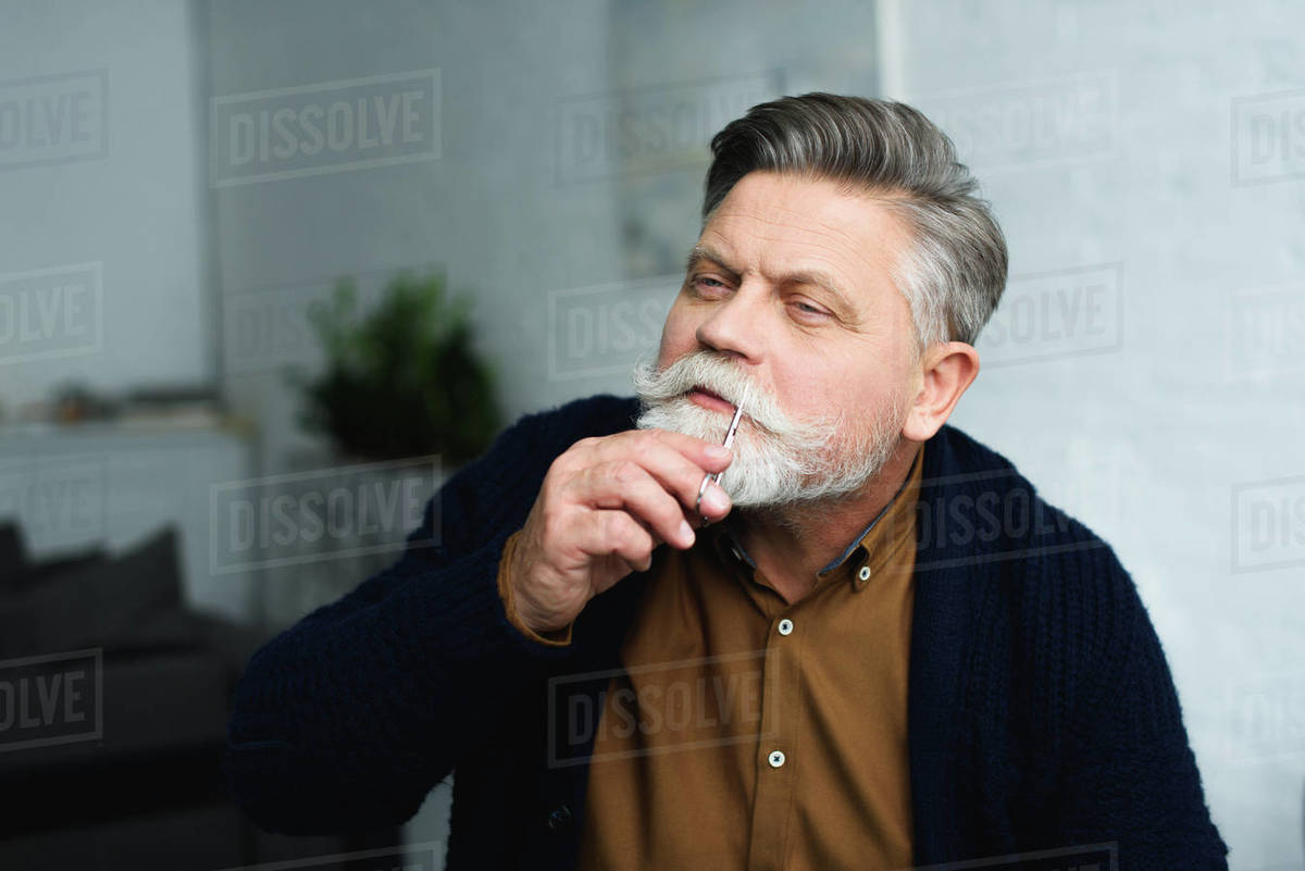 Handsome senior man trimming moustache with scissors at home Stock
