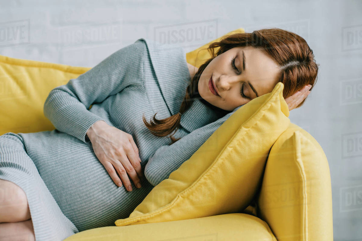 Beautiful pregnant woman sleeping on yellow sofa in living room Stock