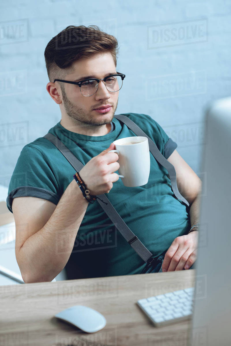 Young man in eyeglasses drinking coffee and working with desktop