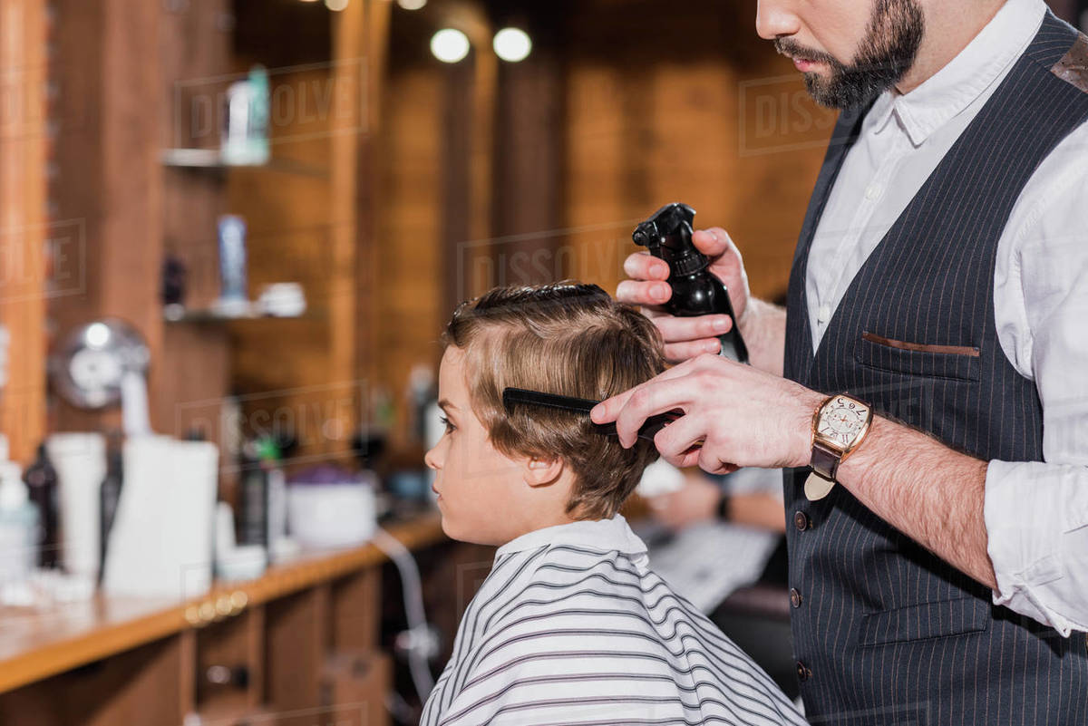 Side view of barber combing and spraying hair of curly kid Stock