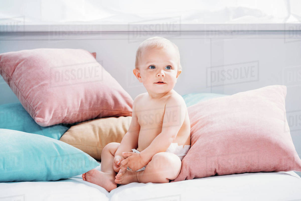 Happy Little Baby Sitting In Bed With Lot Of Pillows And Looking