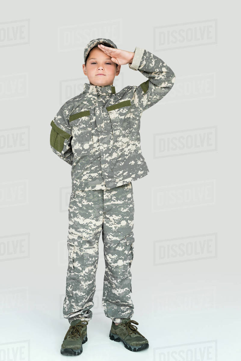 little kid in military uniform looking at camera and saluting on grey
