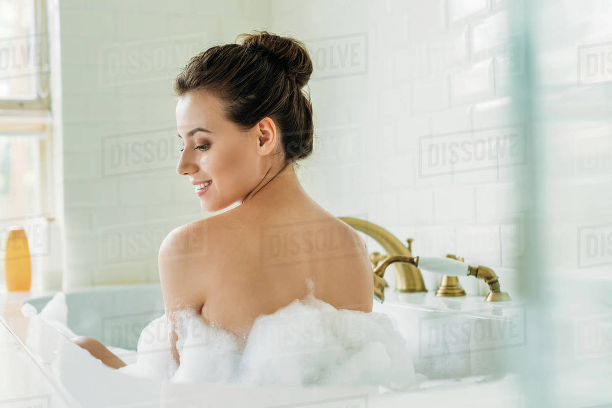 Back view of beautiful smiling naked girl sitting in bathtub with foam