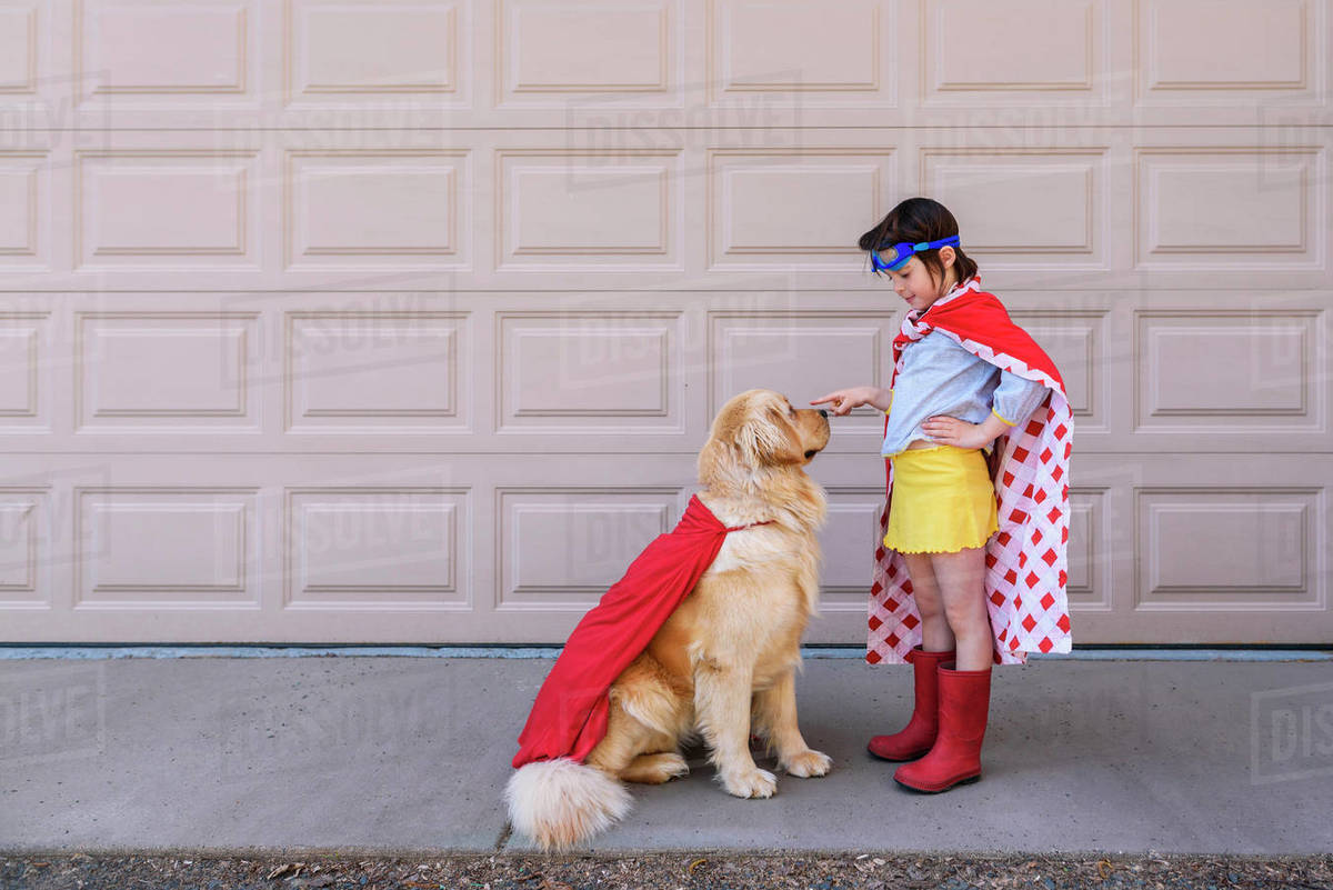 Girl dressed as a superhero standing by the garage with her golden retriever dog Stock Photo