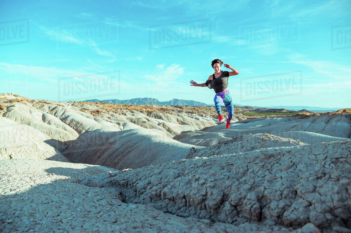Full body active female athlete in sportswear running and jumping over rocky slope of rough desert hilly terrain under blue sky Royalty-free stock photo