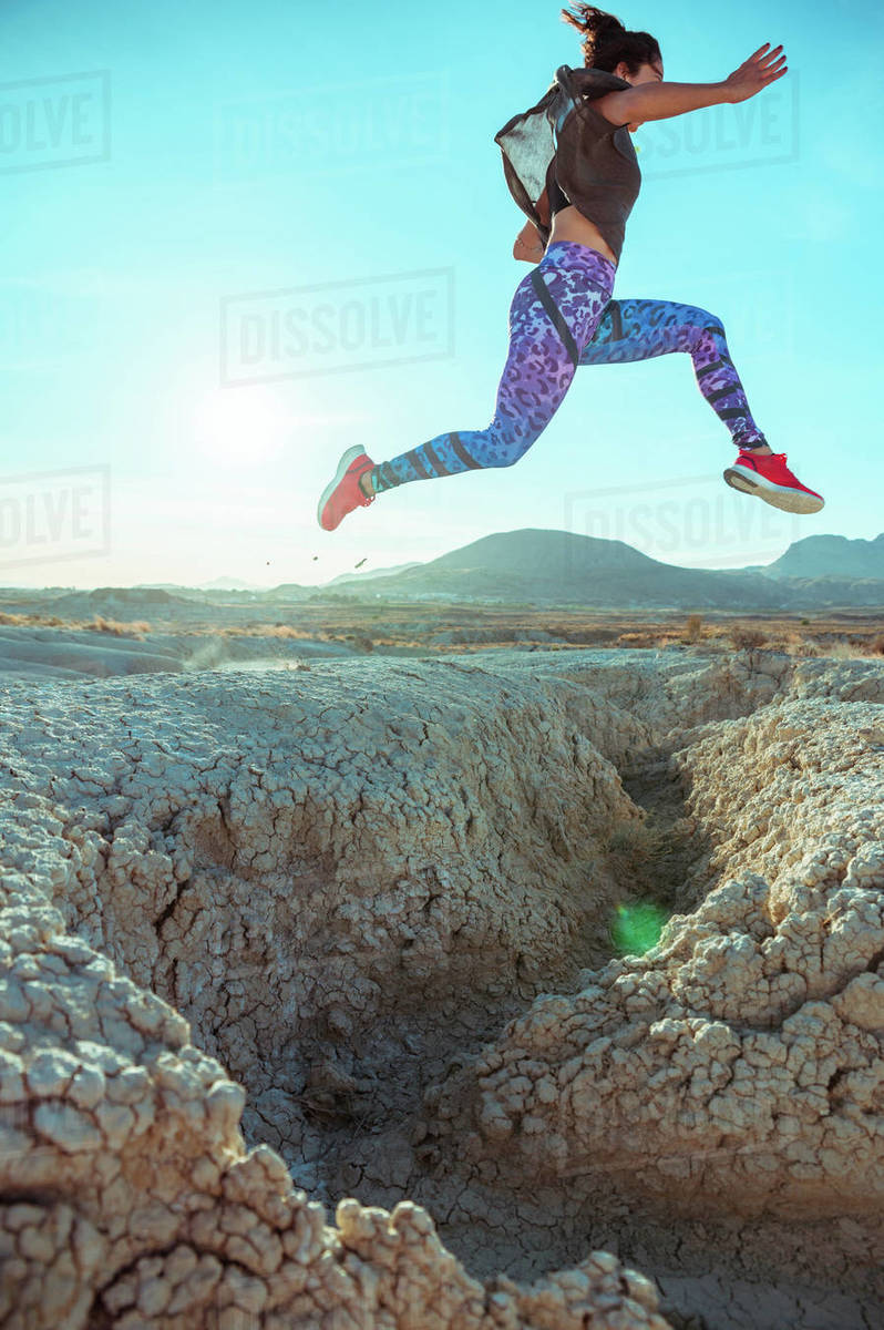 From below full body of active energetic female in sportswear leaping high above sandy slope of desert terrain against blue cloudless sky Royalty-free stock photo