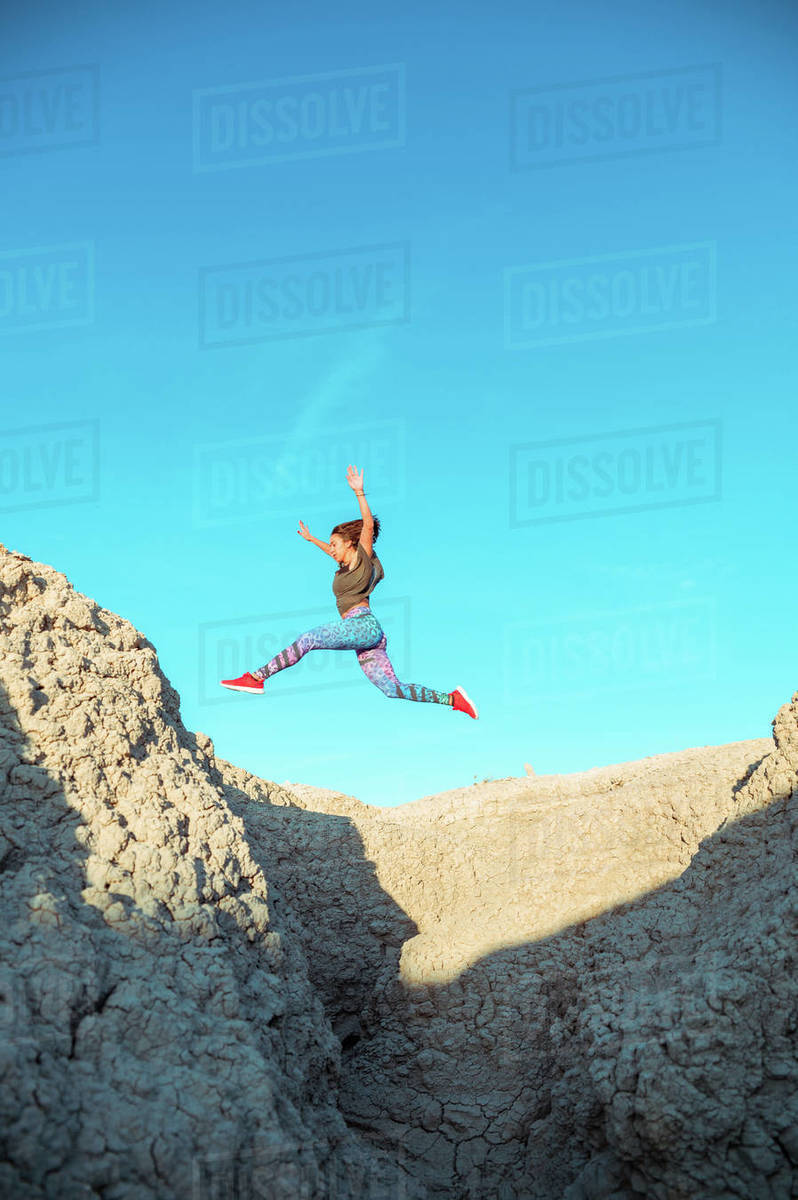 From below full body of active energetic female in sportswear leaping high above sandy slope of desert terrain against blue cloudless sky Royalty-free stock photo