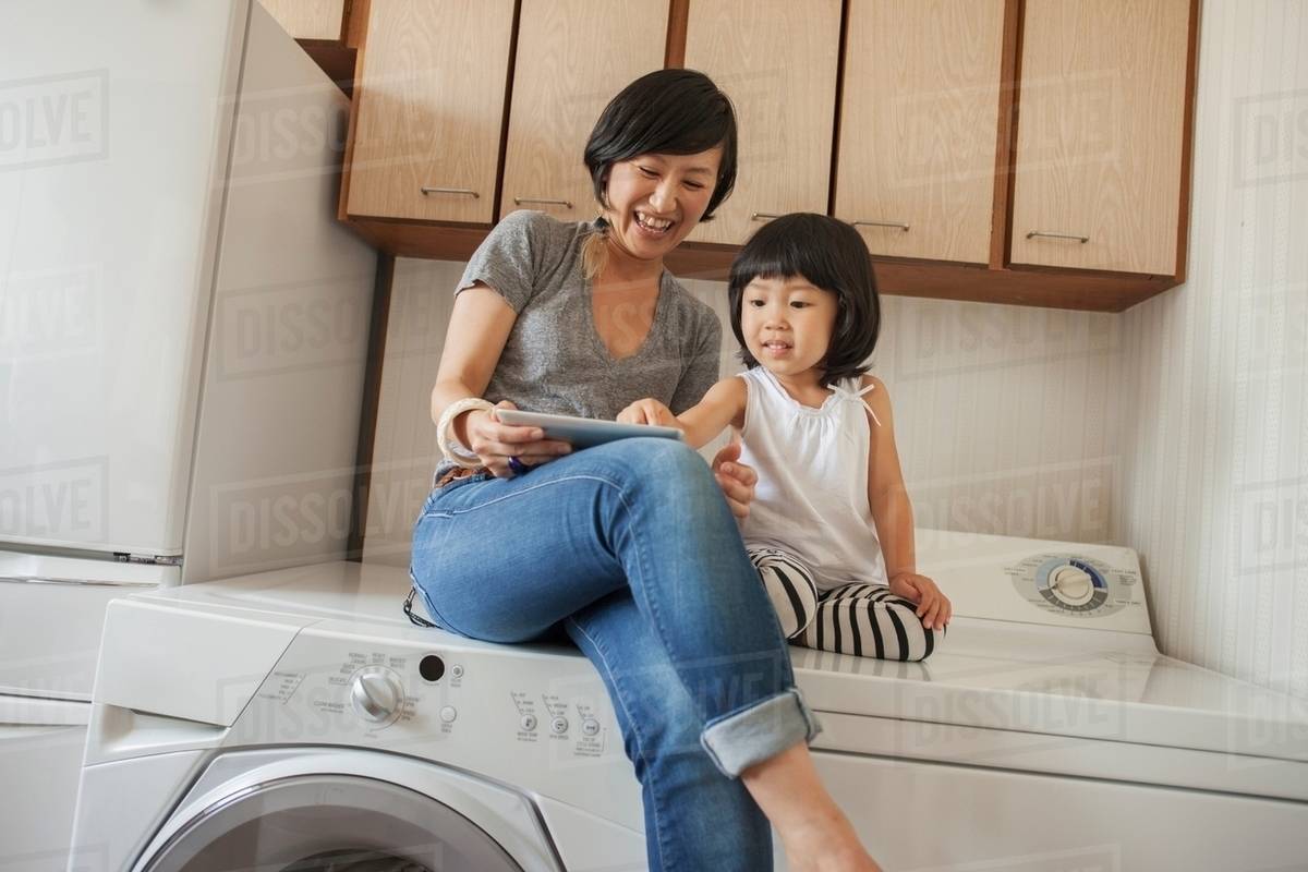 Mother and daughter sitting on washing machine using tablet Stock