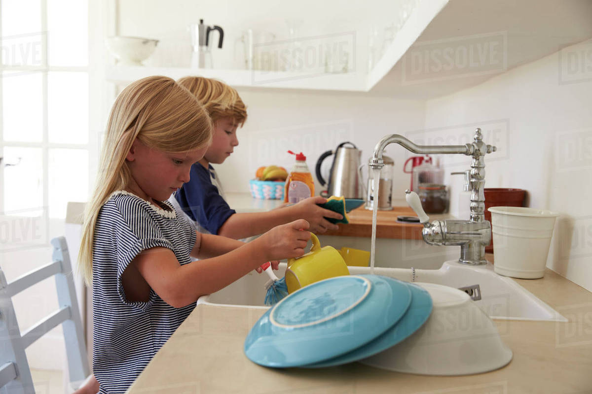 Brother and sister kneeling on chairs to wash up in kitchen Stock