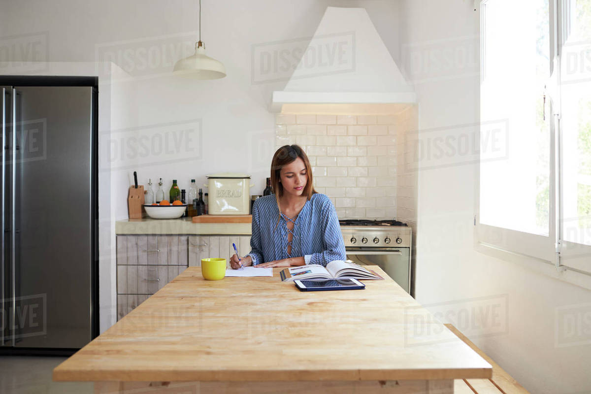 Woman with book and tablet computer writing at kitchen table Stock