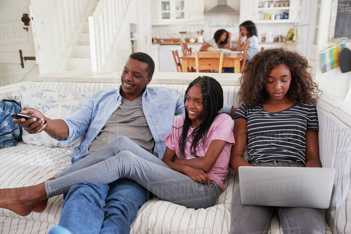 Father Sitting On Sofa Watching Tv With Teenage Daughters Stock