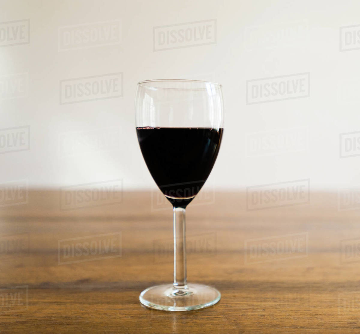 One glass half full of dark red wine standing on wood table against white wall in blurred