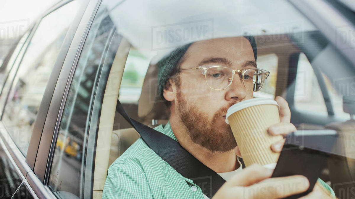 Stylish Young Man Riding in a Car, Sitting on a Passenger Seat Uses Smartphone, types Message, Browses Through Internet, Drinks Coffee. Camera Shot from Outside the Vehicle. Royalty-free stock photo