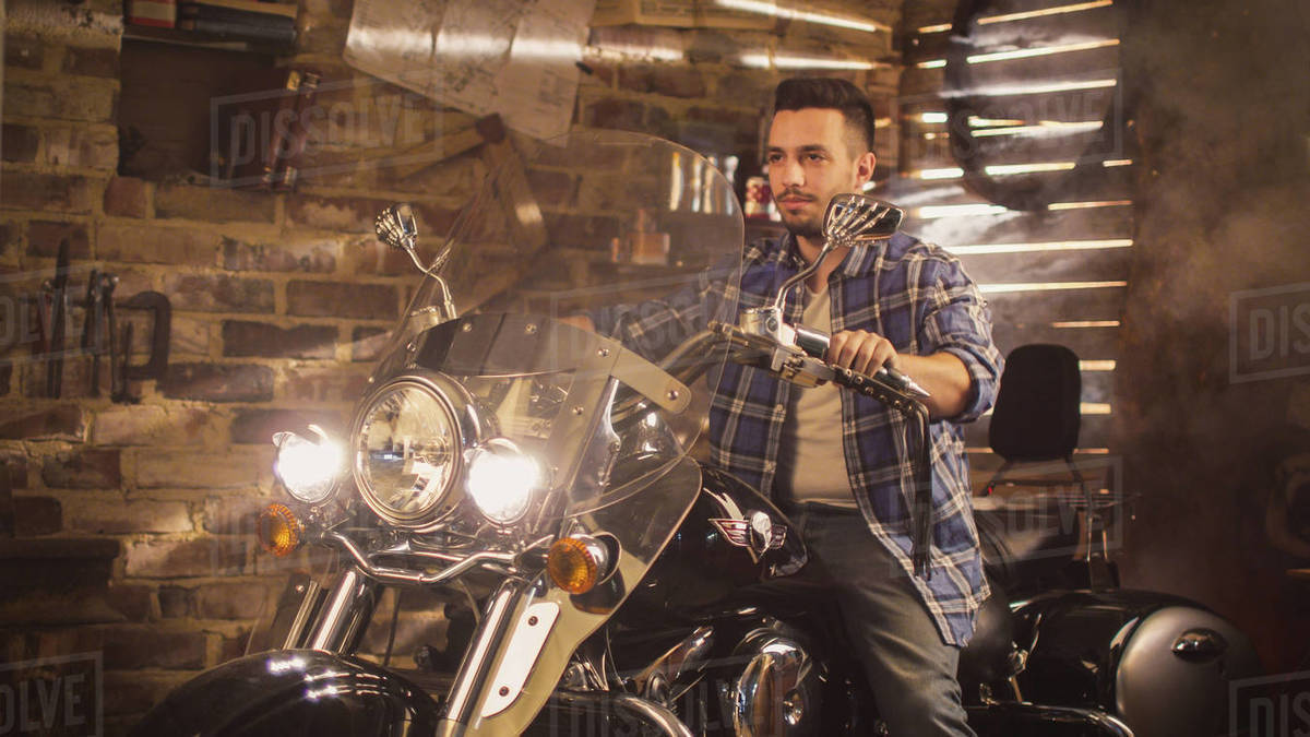 Man started his motorcycle and revs engine in a garage. He is sitting