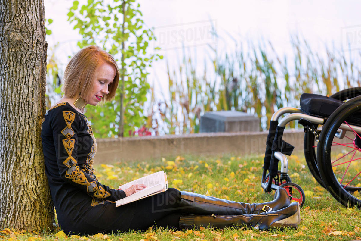 Young disabled woman sitting beside her wheelchair reading a book in a city park in autumn