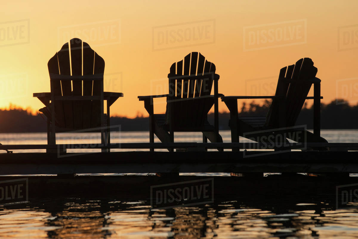 Silhouette of three adirondack chairs on a dock at sunset; Ontario, Canada Stock Photo Dissolve