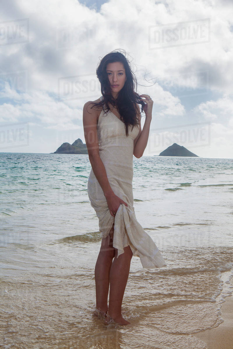 Young woman in white dress on the beach at the water's edge; Kailua, Island of Hawaii, Hawaii, United States of America - Royalty-free Stock Photo | Dissolve Young woman in white dress on the beach at the water's edge; Kailua, Island of Hawaii, Hawaii, United States of America - Royalty-free Stock Photo | Dissolve