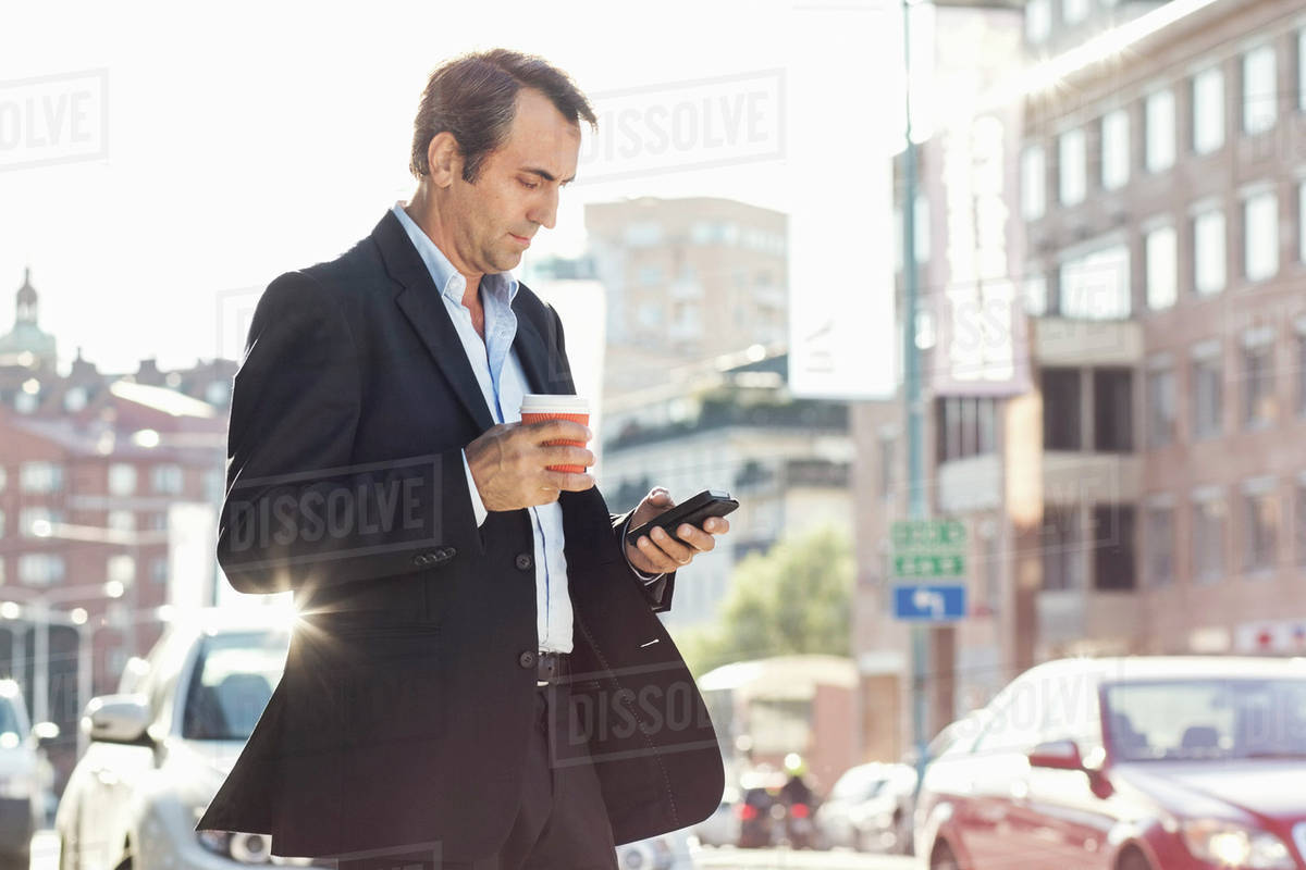 Businessman using mobile phone and holding disposable coffee cup while walking on city street Royalty-free stock photo