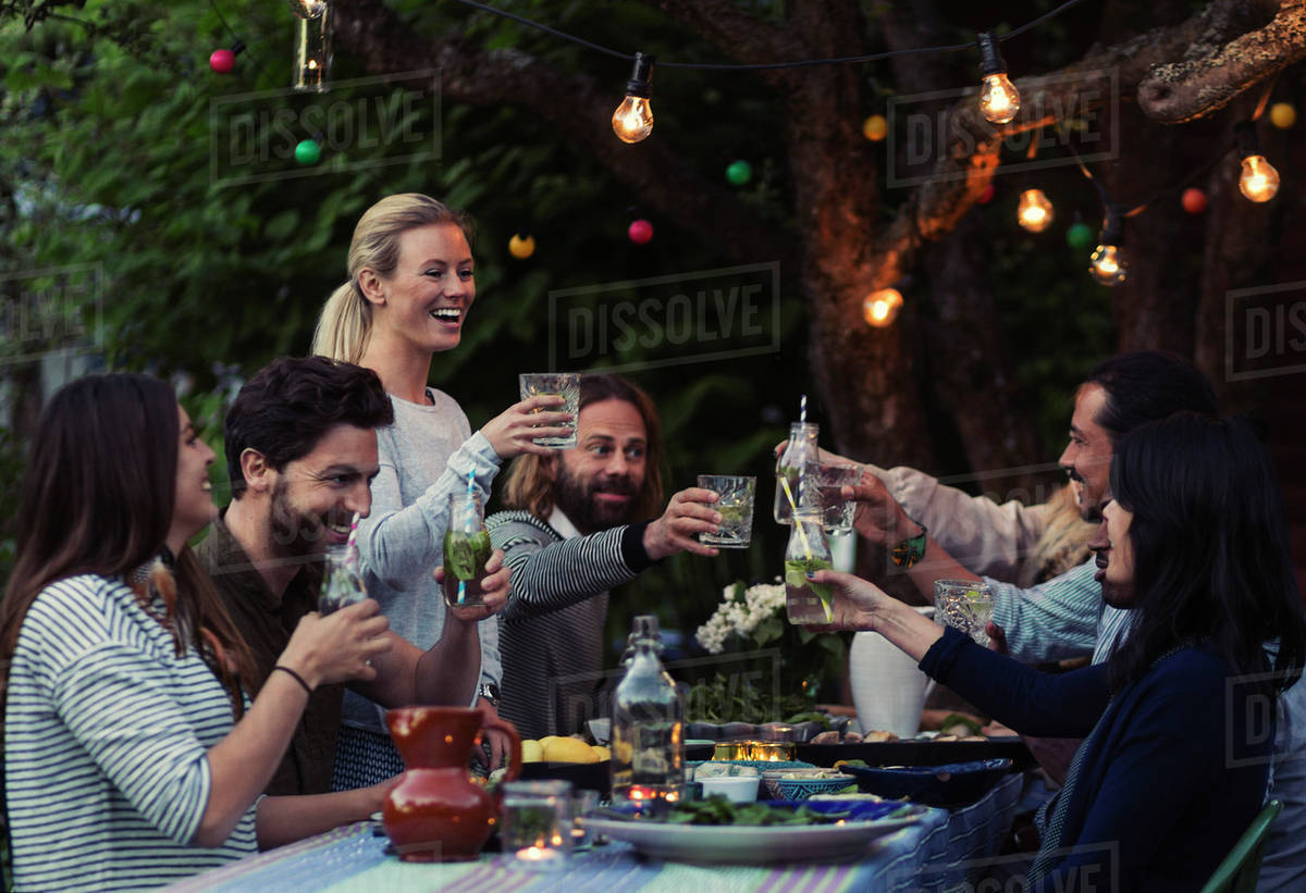 Multi-ethnic friends toasting drinks at dinner table in yard - Stock