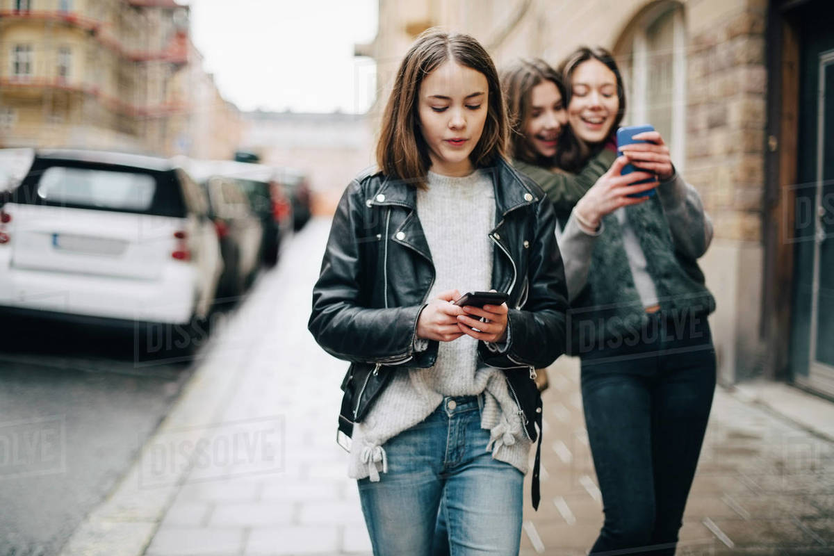 Female friends using mobile phone while walking on sidewalk in city