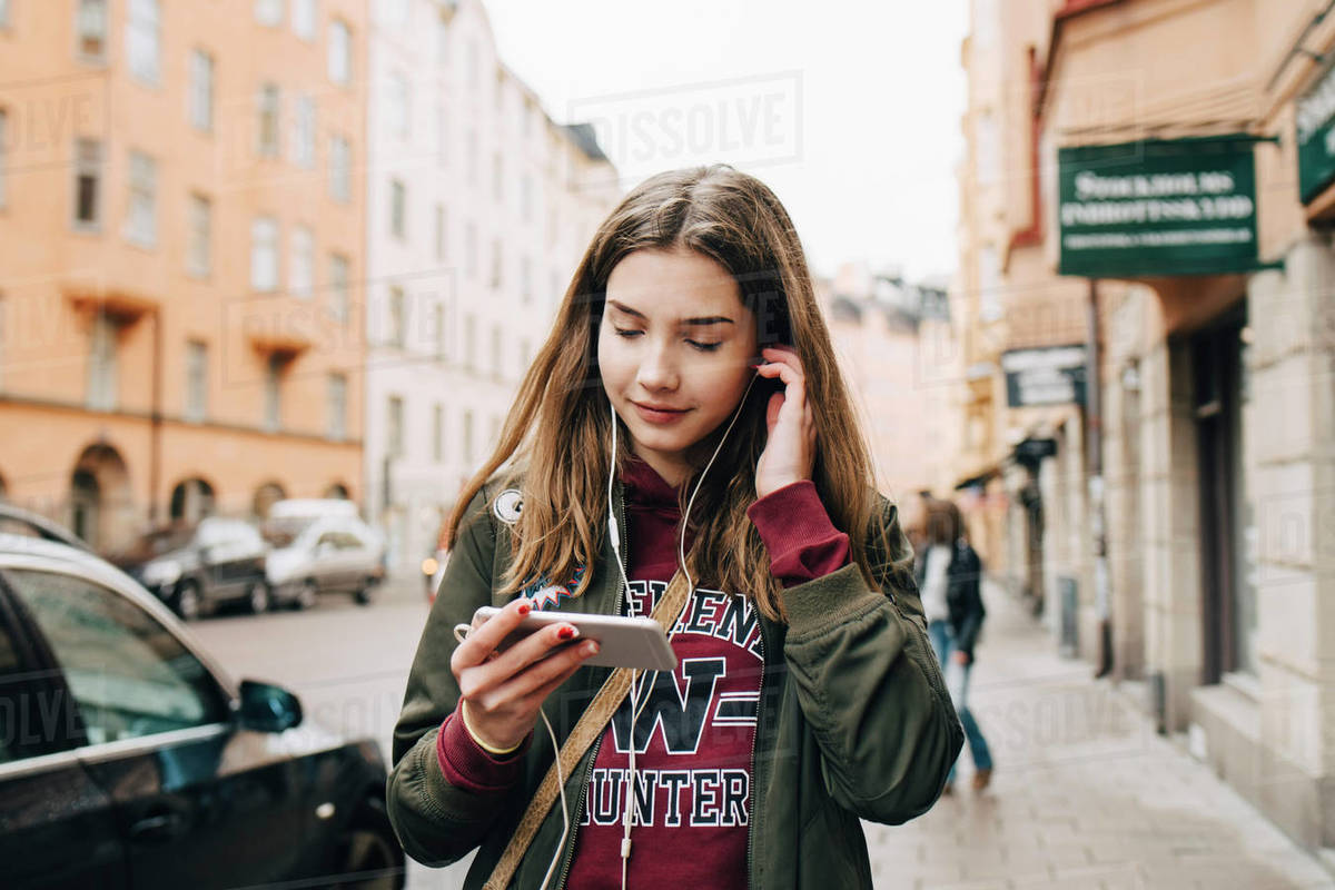 Smiling girl listening music through smart phone while walking on