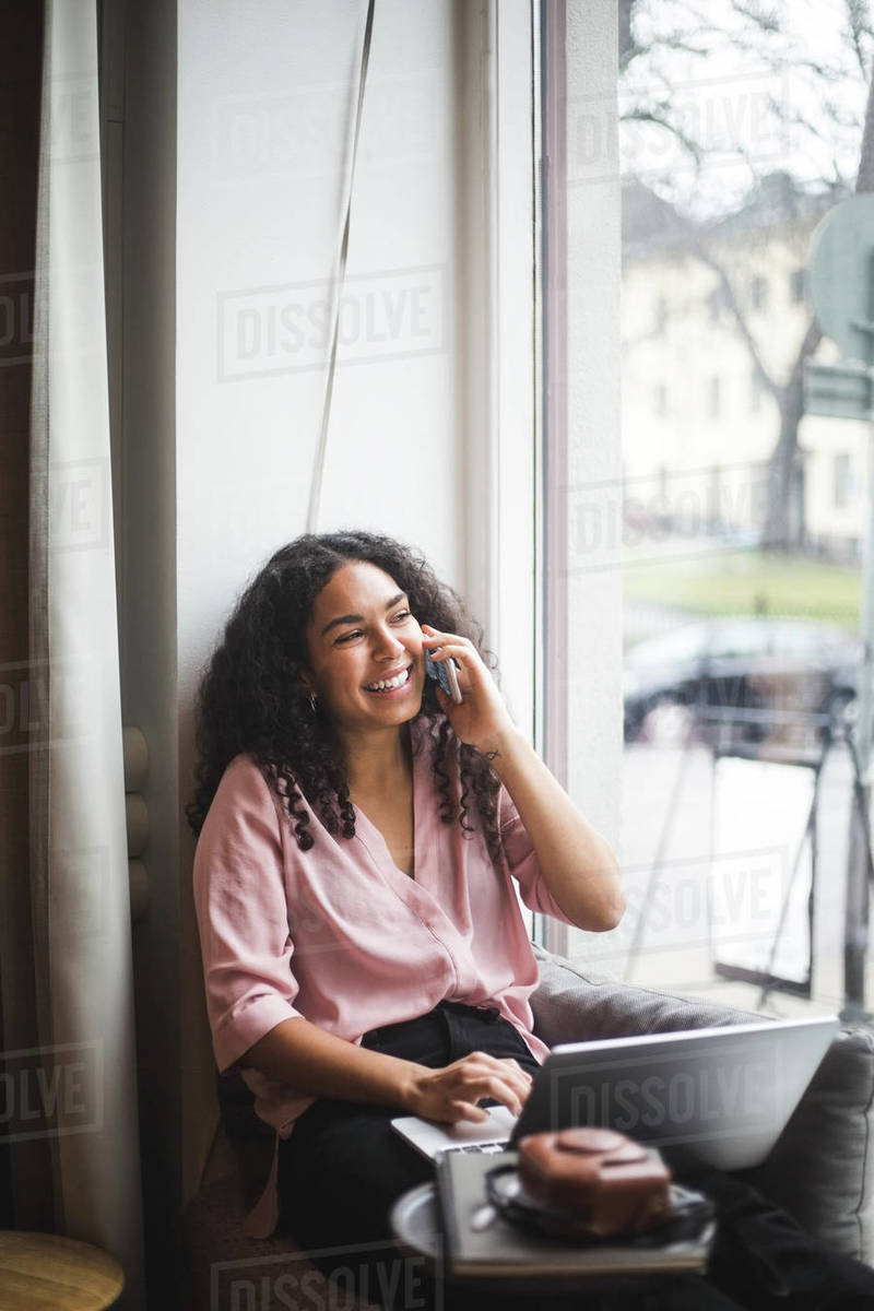 Smiling young female entrepreneur talking through smart phone sitting on window sill in office Royalty-free stock photo