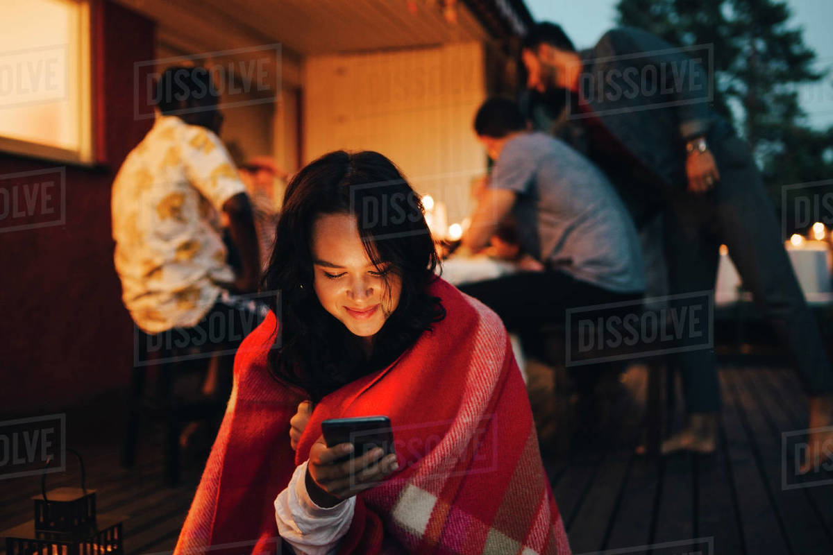 Smiling young woman using mobile phone while friends in background during dinner party Royalty-free stock photo