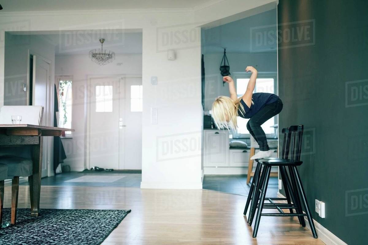 Side view of girl jumping from chair in living room at home Stock